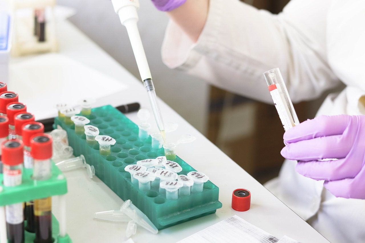 Close up of a gloved hand holding a dropping, filling a test tube with liquid. The test tube sits in a tray of tubes on a desk, and another full tray sits nearby.