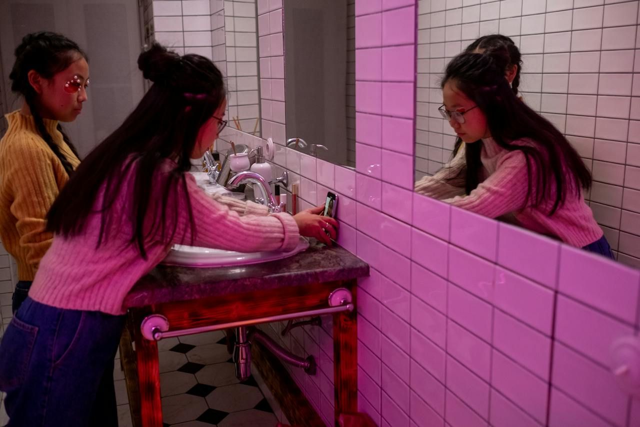 Two young people in a neon pink bathroom prop a phone up against the wall on the sink