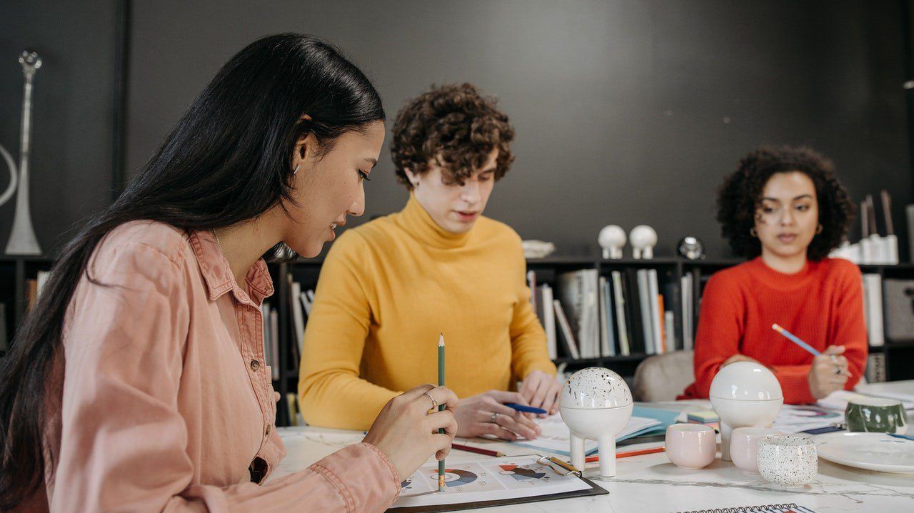 Three people sitting around a desk, holding pencils and working on documents together.