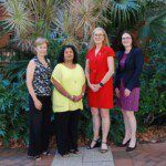 Four women stand in together in front of some bushes and a building