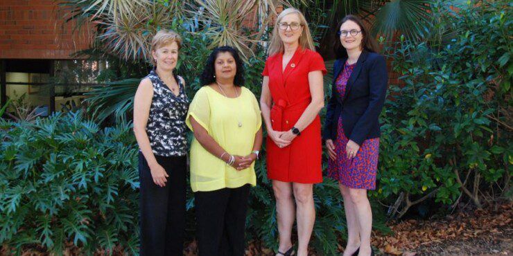 Four women stand in together in front of some bushes and a building