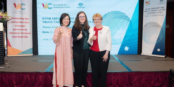 Three women stand together giving thumbs up in front of a display screen showing the logo for Vietnam Australia Centre