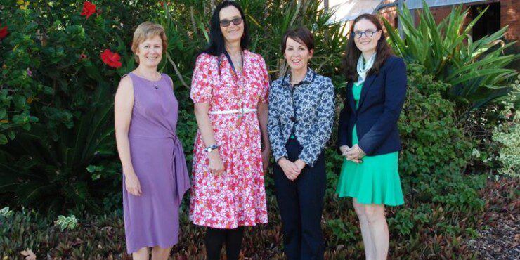 Four women stand together smiling at the camera in front of a low brick wall and greenery