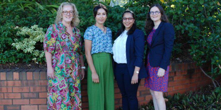 Four women stand together smiling at the camera in front of a low brick wall and greenery