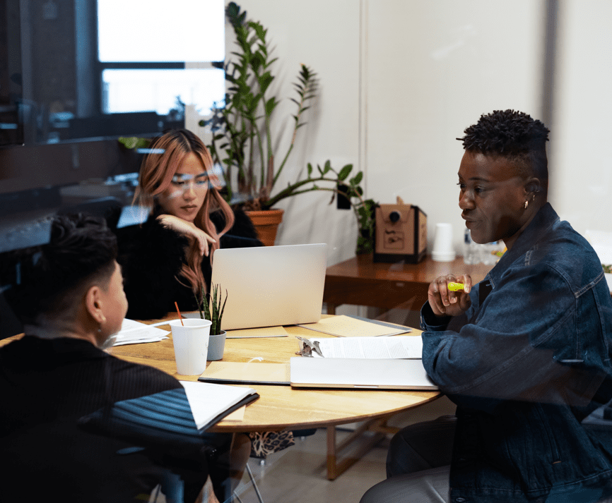 Three people sit around a table with laptops talking