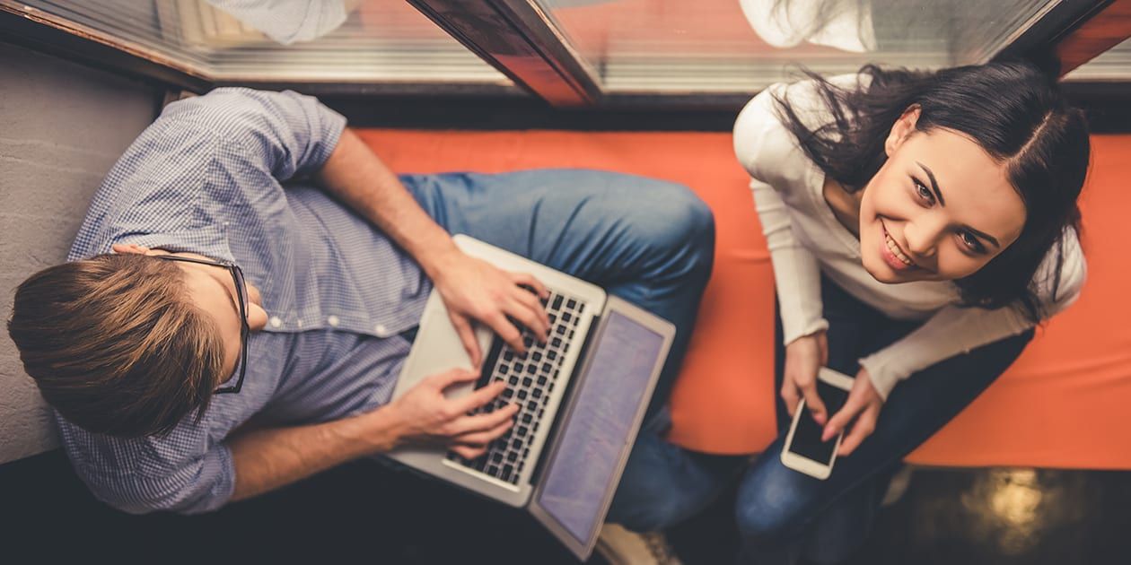 A male students sitting on bench, looking at a laptop next to a females student, looking up at camera and smiling