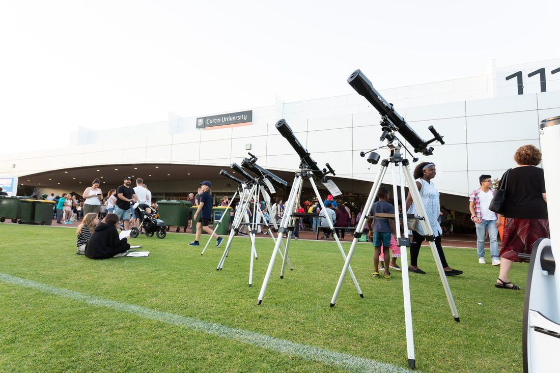 Line of telescopes outside Curtin Stadium on the grass pointing at the sky. There are people moving around the telescopes.