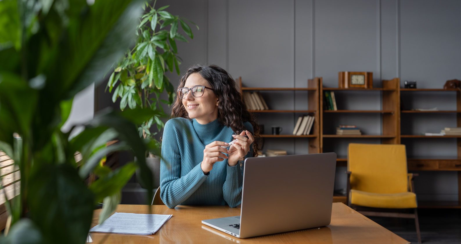 A student smiling while looking out a window. She sits at a desk with a laptop in front of her.