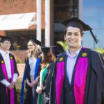 Young male graduate in graduation regalia and smiling and standing in front of a small group of graduates