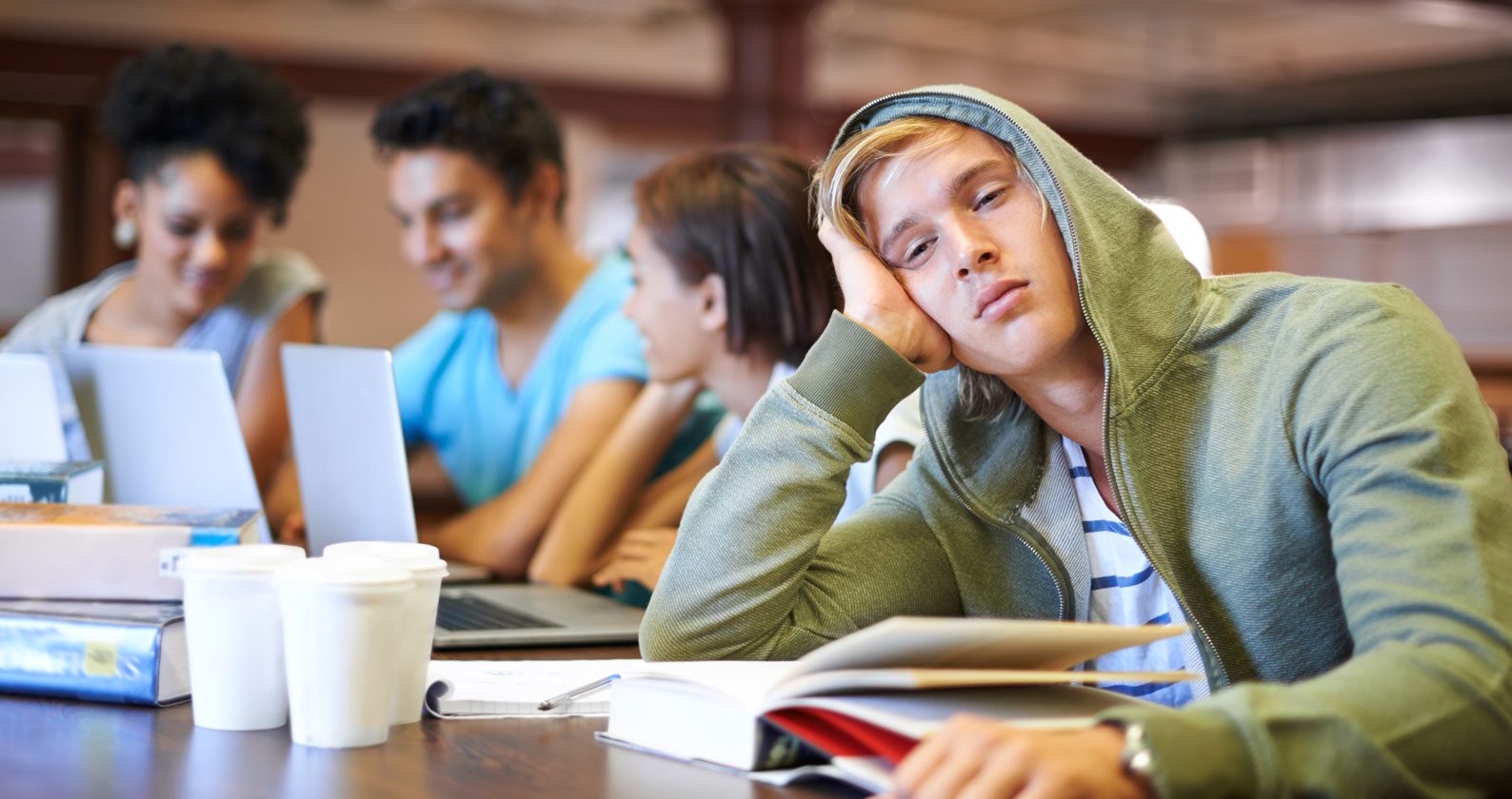 Student sitting at table surrounded by books and empty coffee cups.