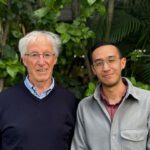 David and Bruno smile at the camera. They stand in front of a wall of plants.