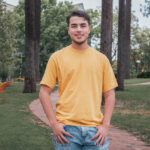 Andrew smiles wearing a yellow t-shirt in Henderson Court. He hands his hands in the pockets of his jeans, and has a big smile on his face.