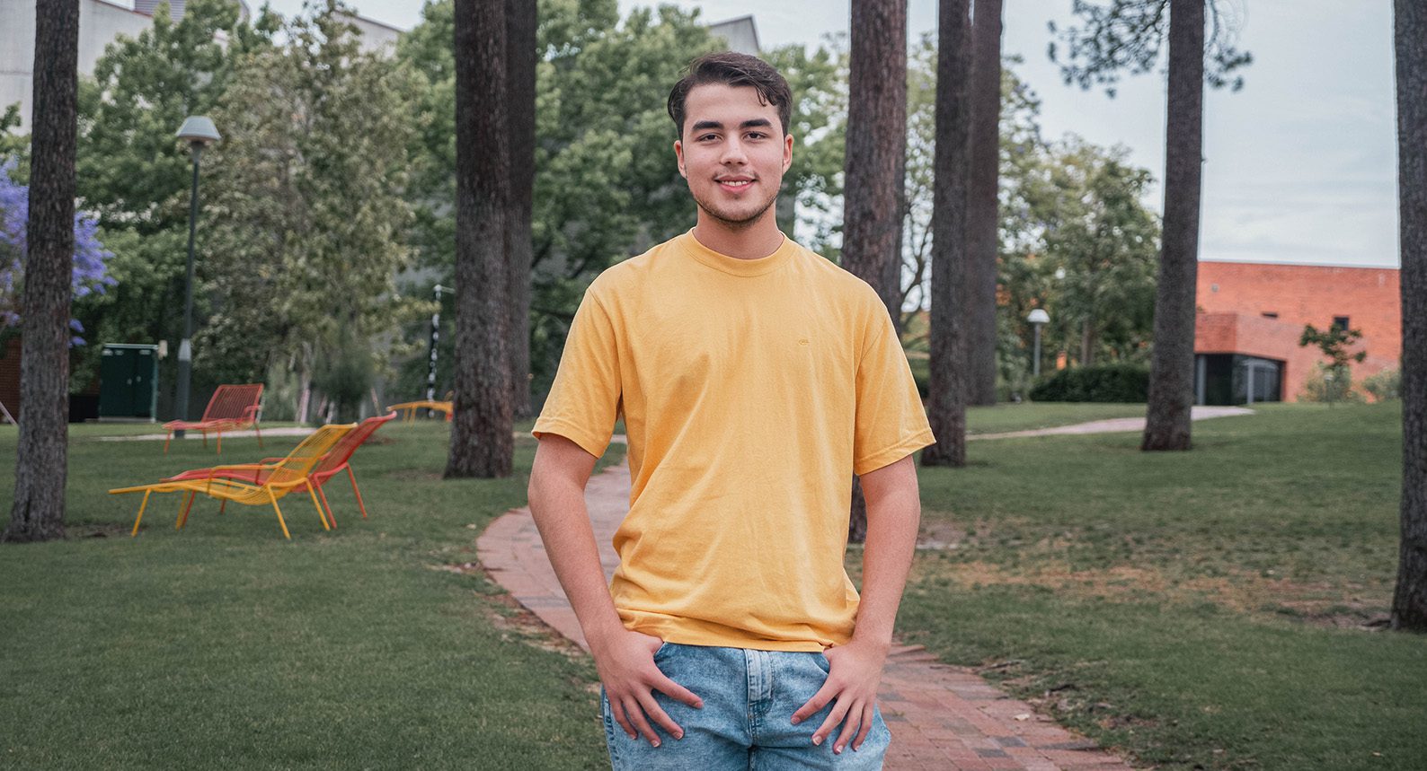 Andrew smiles wearing a yellow t-shirt in Henderson Court. He hands his hands in the pockets of his jeans, and has a big smile on his face.