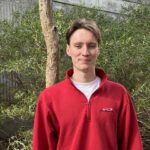 Pierce stands smiling at the camera, he wears a red jumper with a white t-shirt underneath. He is standing outside in front of foliage and trees.