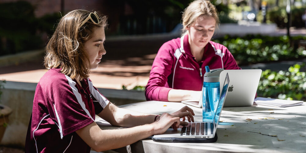 2 students on their laptops, sitting at a table outside.