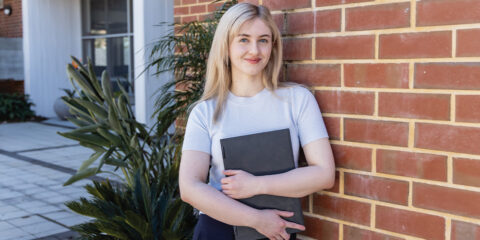 Student leaning on a red brick wall holding a notebook