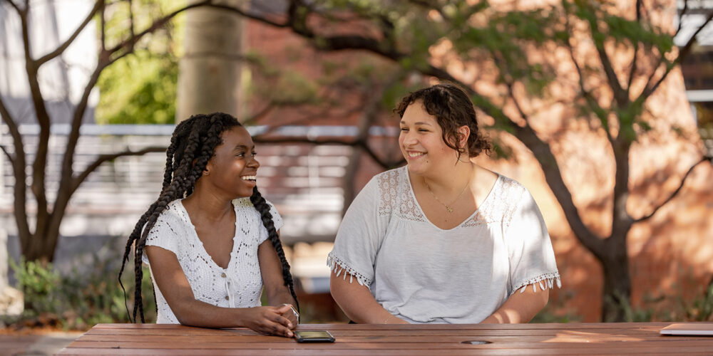 Two students smiling at each other while sitting on a wooden table