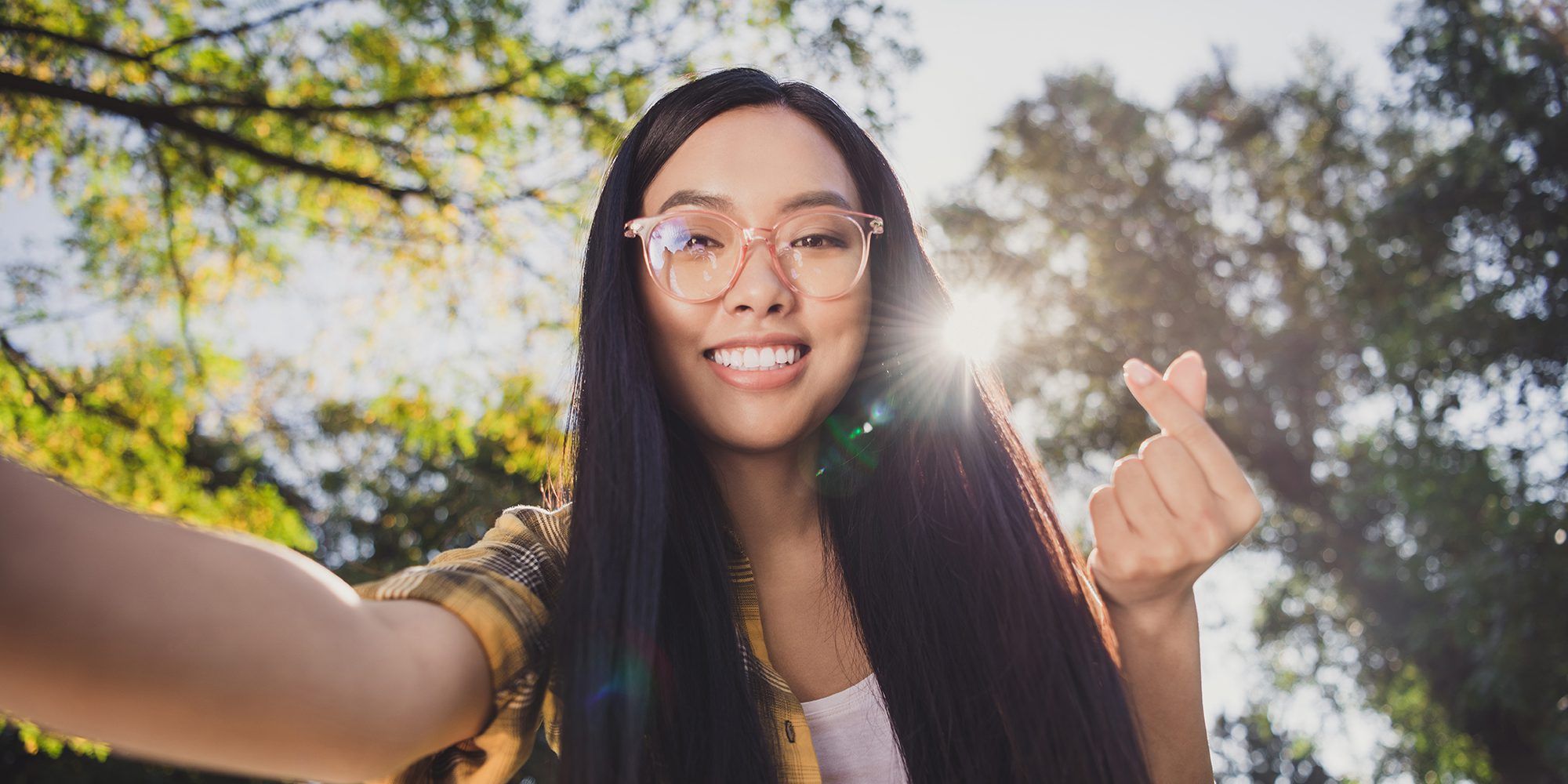 Photo of pretty cute young woman dressed plaid shirt spectacles recording self video showing korean love sign outside countryside nature.