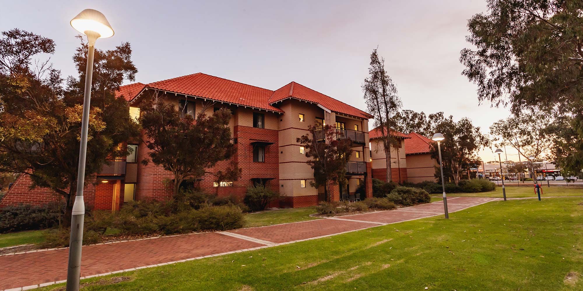 Outside one of the student accomodation buildings at Kurrajong Village. The building is three storeys high and brick with grass and trees in front.