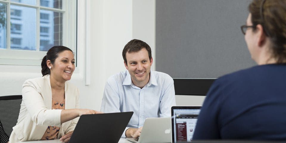 Two young professionals sitting at a desk with a laptop and speaking with another student