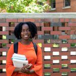 Female student holding papers and books , standing in front of a wall