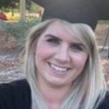 A young woman with light skin and blonde hair wears a graduation cap and gown.