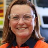 A woman with white skin and red hair smiles. She is wearing safety glasses and a high vis shirt.