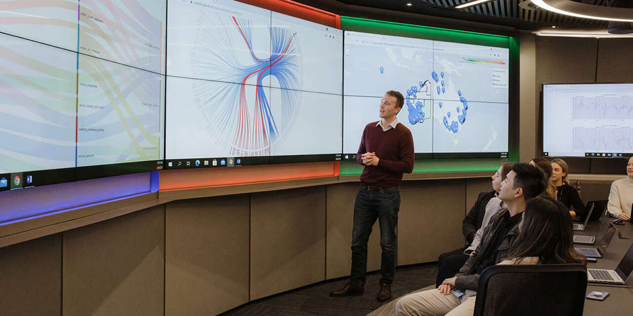 Man dressed in maroon jumper analysing data on big screen in front of class of students.