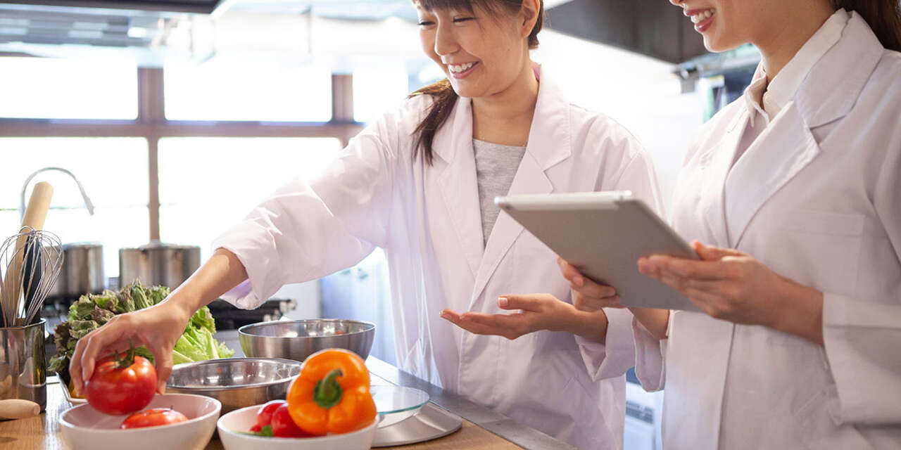 Two women smiling in white laboratory coats as they examine fruits and vegetables.