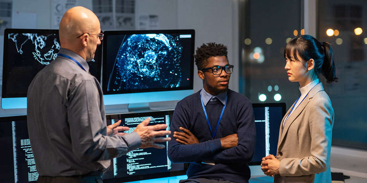 Three intercultural colleagues in formalwear discussing working points at meeting by their workplace in front of computers with data and digitised map.