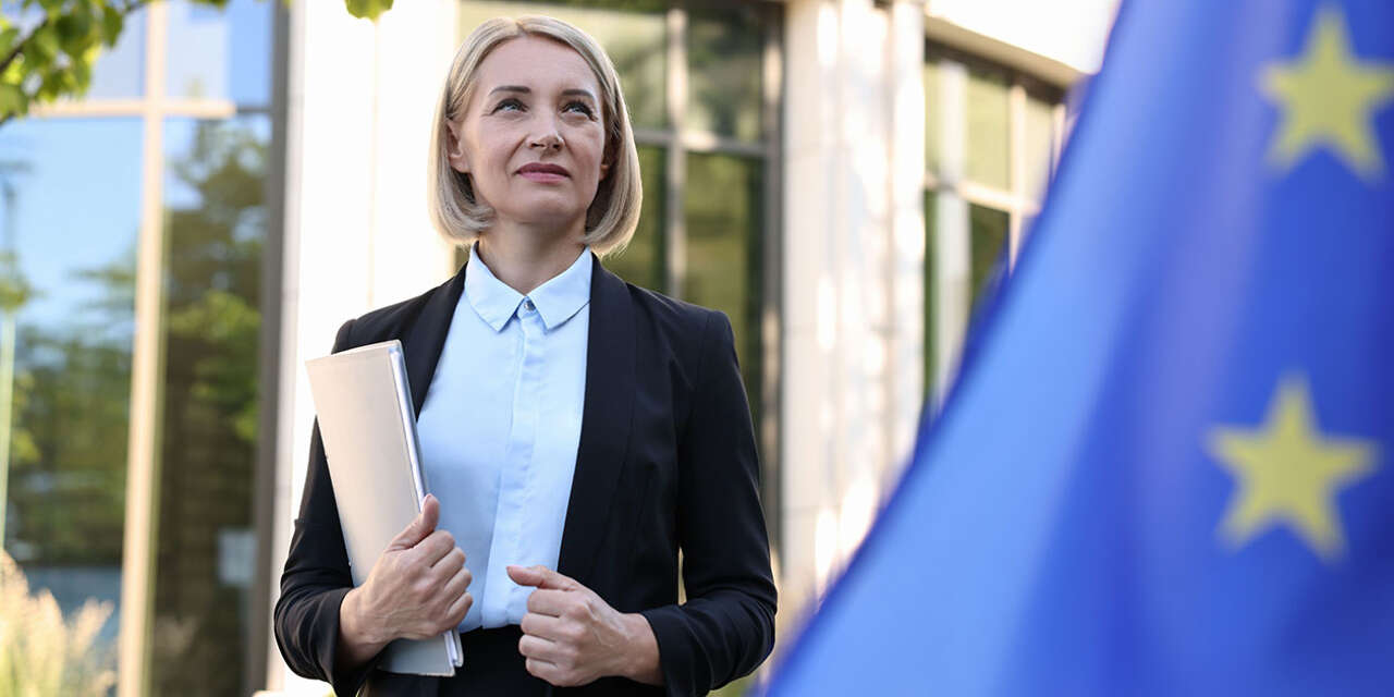 Woman in business attire stands behind a flag looking hopeful with fists loosely clenched.